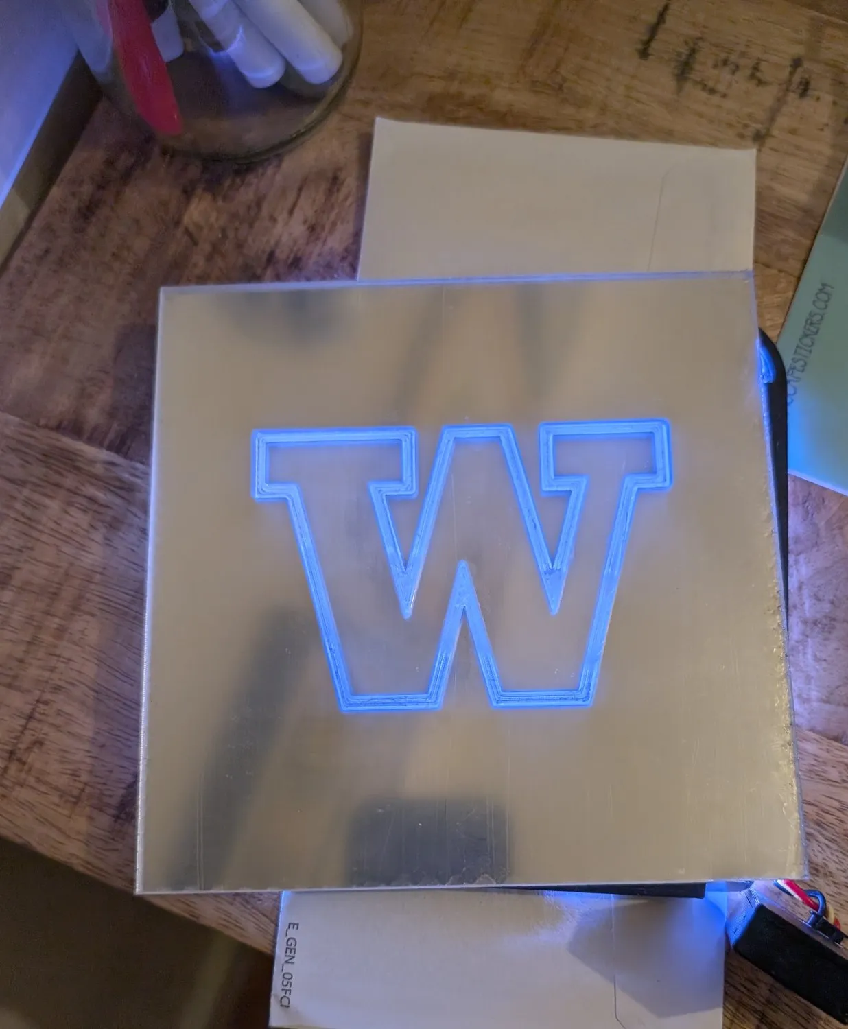 A custom mirror tile on the workbench, the University of Washington W laser-cut through it and glowing blue